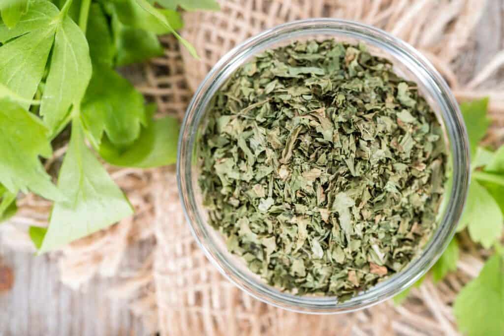 An open jar sits on a table as seen from above. The jar is full of dried herbal leaves. Next to the jar sits a stem of fresh lovage leaves.