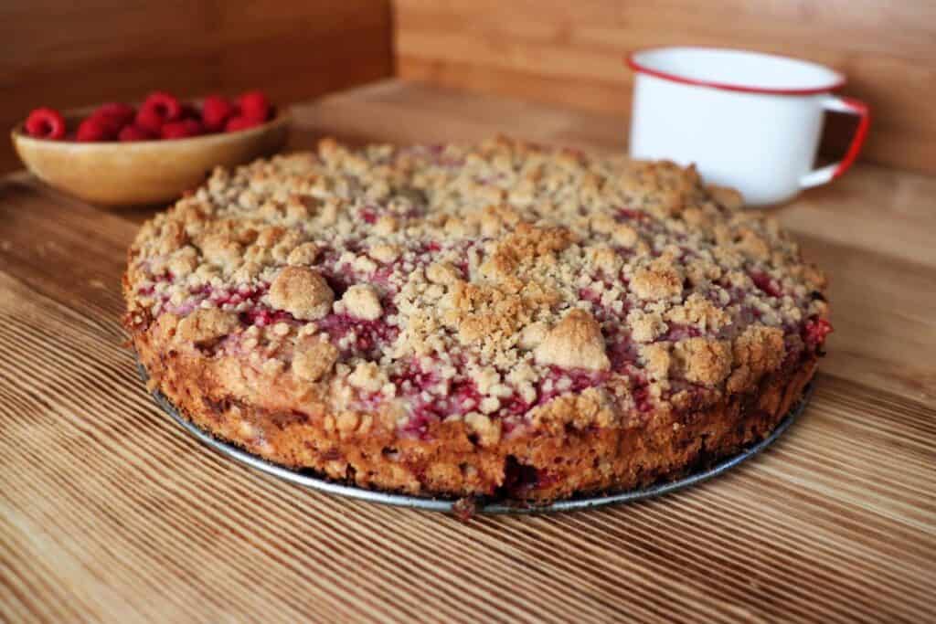 A coffee cake with crumb topping sits on a board. In the background is a white coffee cup and a wooden bowl full of fresh raspberries.