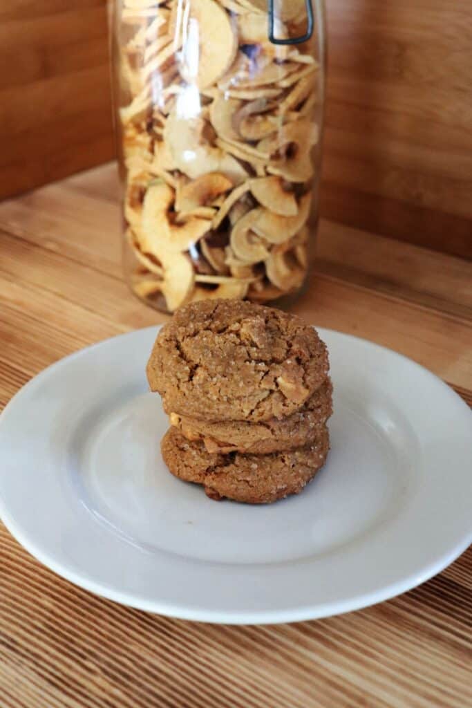 3 molasses ginger cookies with bits of dried apples showing are stacked on a white plate. A jar of dried apple rings sits behind the plate.