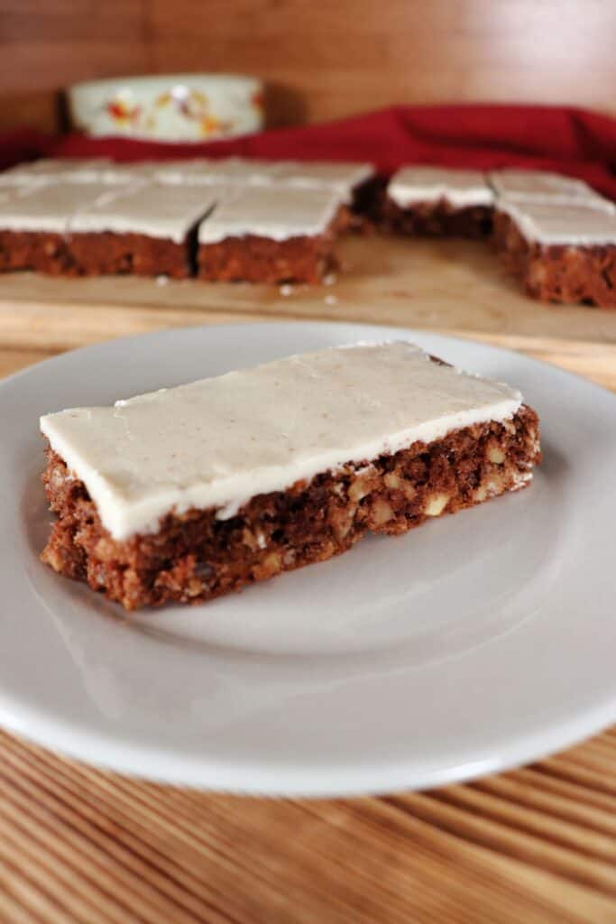 An apple oatmeal bar with thin caramel frosting sits on a plate. A platter with more bars can be seen in the background.