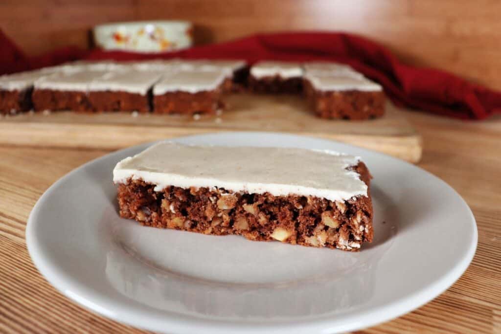 An apple oatmeal bar with thin caramel frosting sits on a plate. A platter with more bars can be seen in the background.