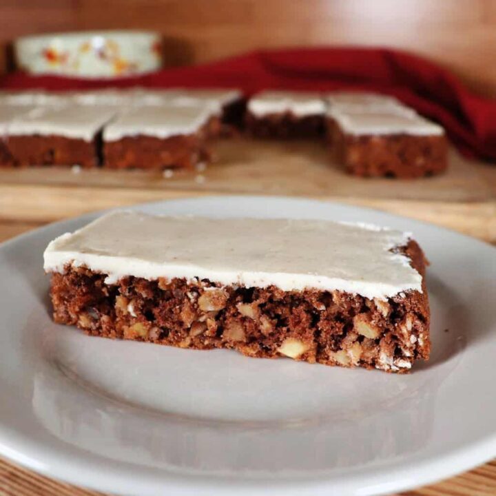 An apple oatmeal bar with thin caramel frosting sits on a plate. A platter with more bars can be seen in the background.