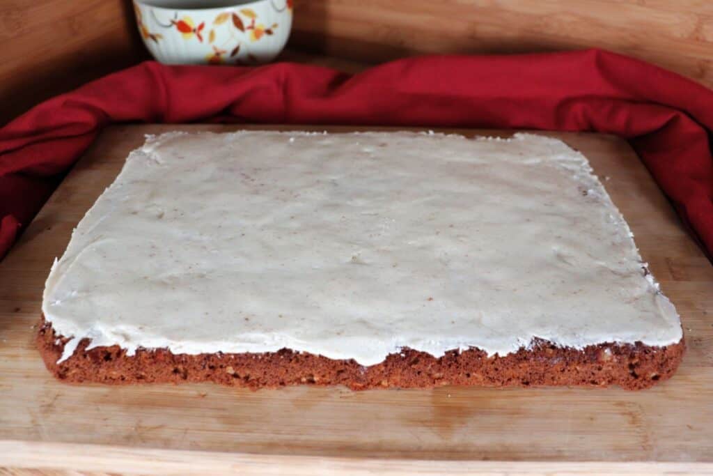 Frosted apple oat bars sit uncut on a cutting board. A red cloth draped behind the board. 