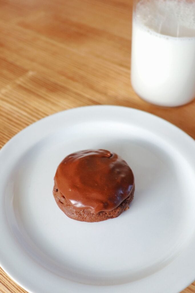 A frosted chocolate cookie sits on a plate with a glass of milk next to it. 