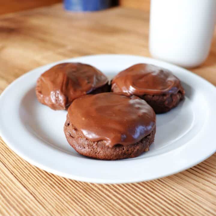 3 frosted chocolate potato cookies sit on a plate with a glass of milk behind it.