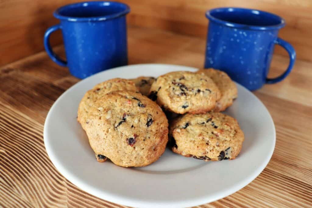 A plate of cookies with raisins and nuts visible sits in front of two blue coffee cups on a table. 