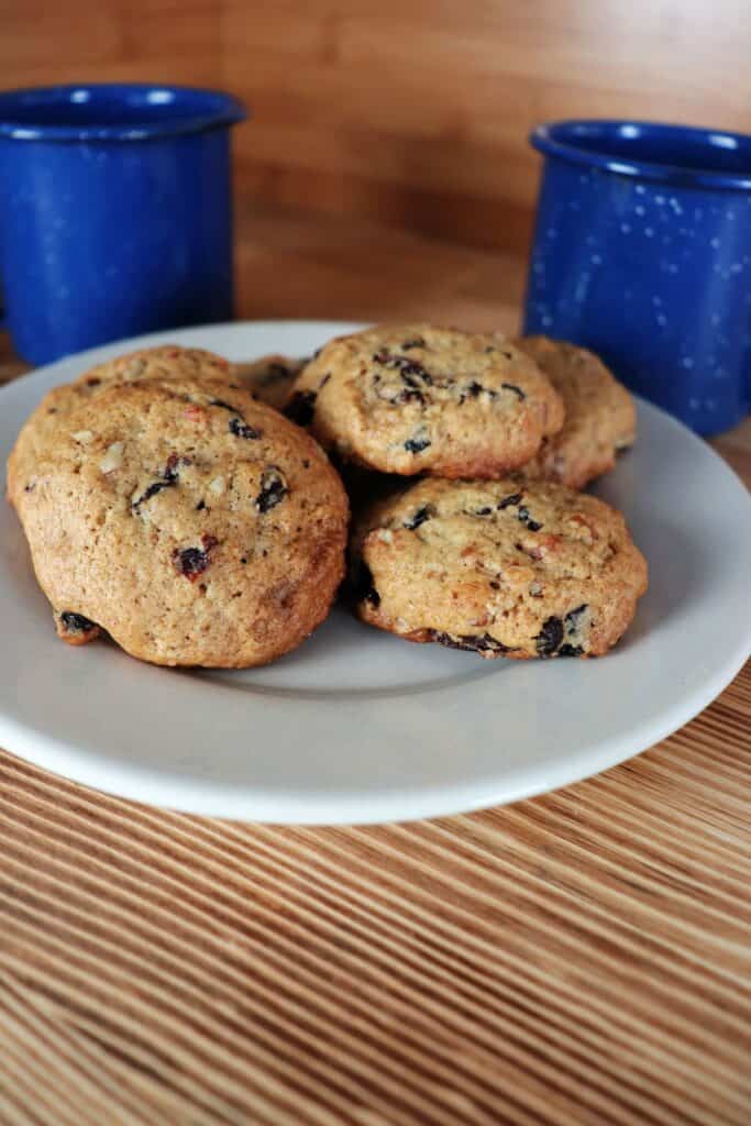A plate of cookies with raisins and nuts visible sits in front of two blue coffee cups on a table. 