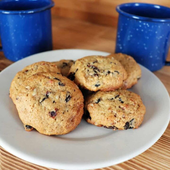 Cookies with raisins and nuts visible sit on a plate with blue coffee cups behind it on a the table.