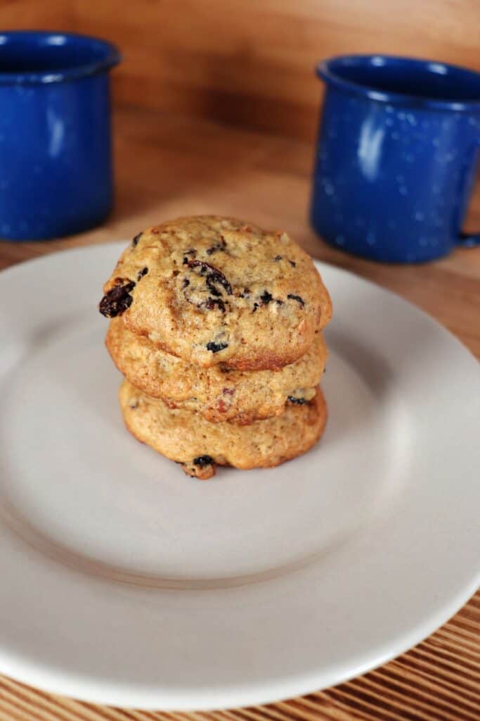 3 cookies with raisins and nuts sit stacked on a plate, blue coffee cups behind it. 