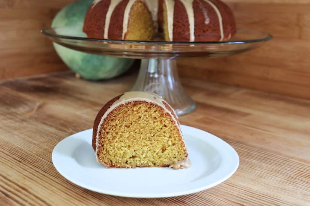 A slice of coffee glazed pumpkin spice bundt cake sits on a plate. Behind it is a glass cake plate on a pedestal with the remaining cake and a blue hubbard squash. T