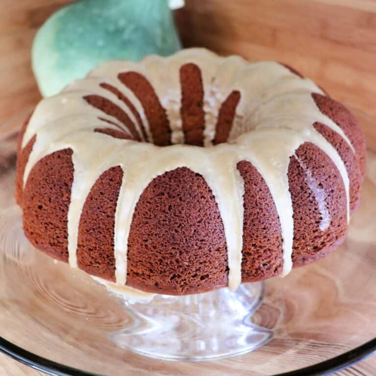 A whole pumpkin spice cake with coffee glaze sits on a glass cake plate. A blue hubbard squash in the background.