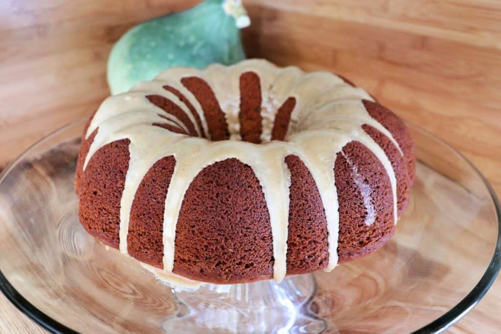 A whole pumpkin bundt cake with coffee frosting sits on a glass cake plate. A blue hubbard squash sits in the background.