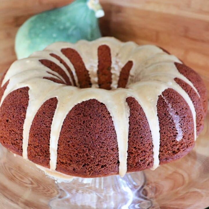 A whole pumpkin bundt cake with coffee frosting sits on a glass cake plate. A blue hubbard squash sits in the background.