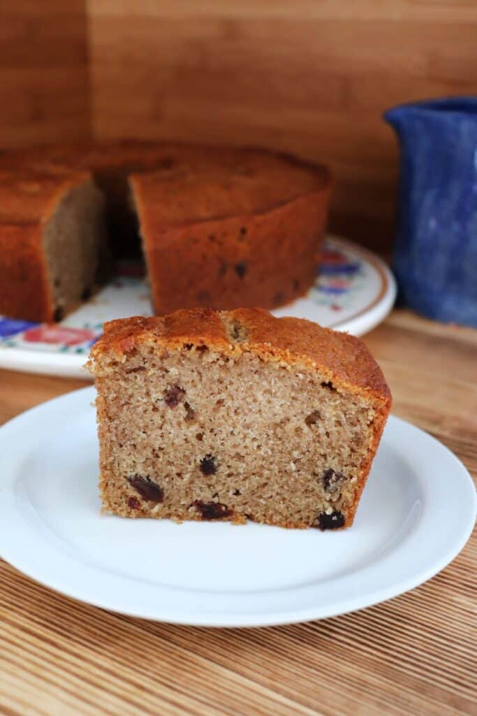 A slice of raisin spice cake sits on a plate. The remaining cake and a blue pitcher sit in the background.