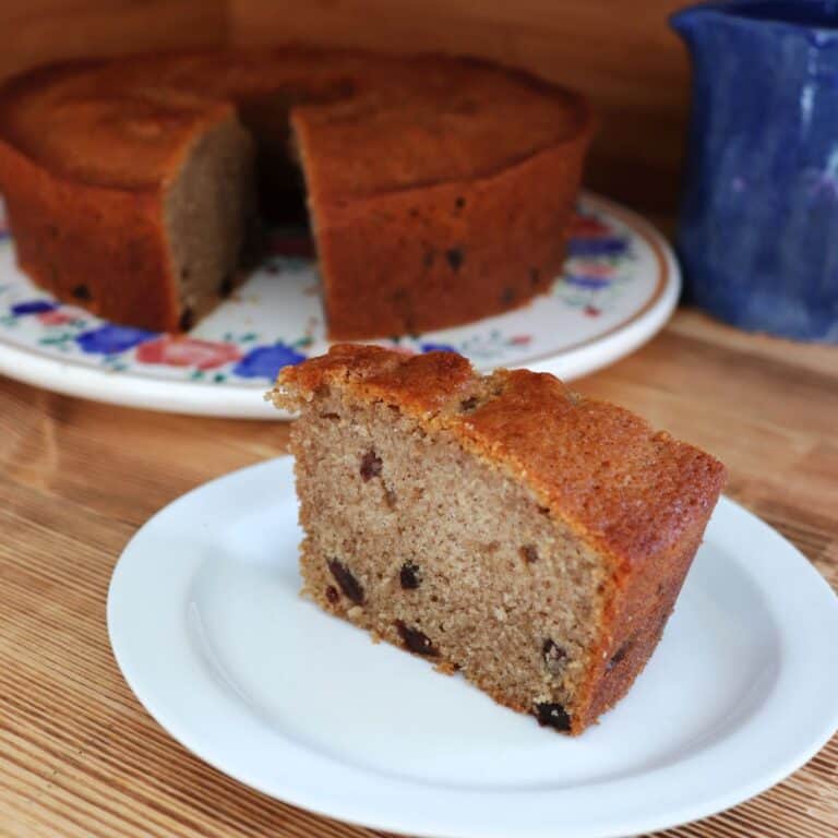 A slice of raisin spice cake sits ona plate. Behind it is the remaining cake and a blue clay pitcher.