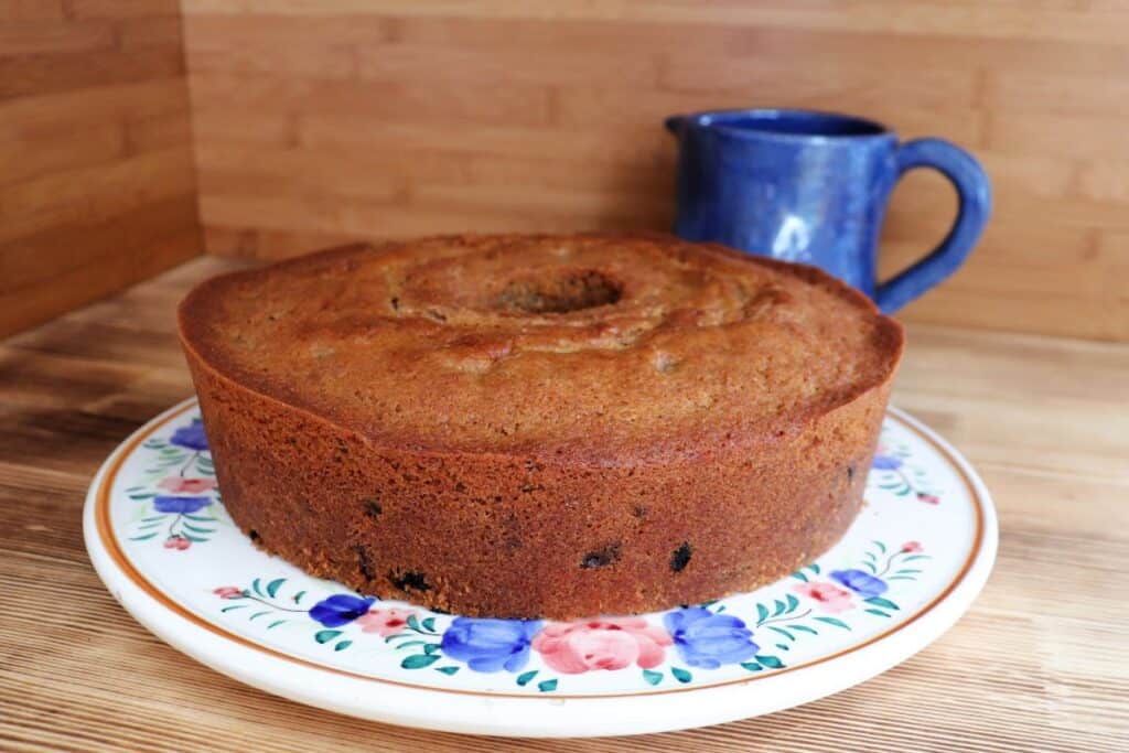A whole raisin space cake sits on a flowered cake plate with a blue pitcher behind it.