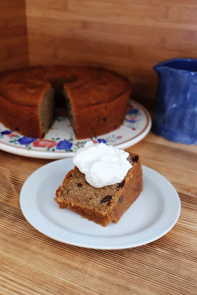 A slice of cake with whipped cream on top sits on a plate. The remaining cake sits in the background with a blue pitcher.